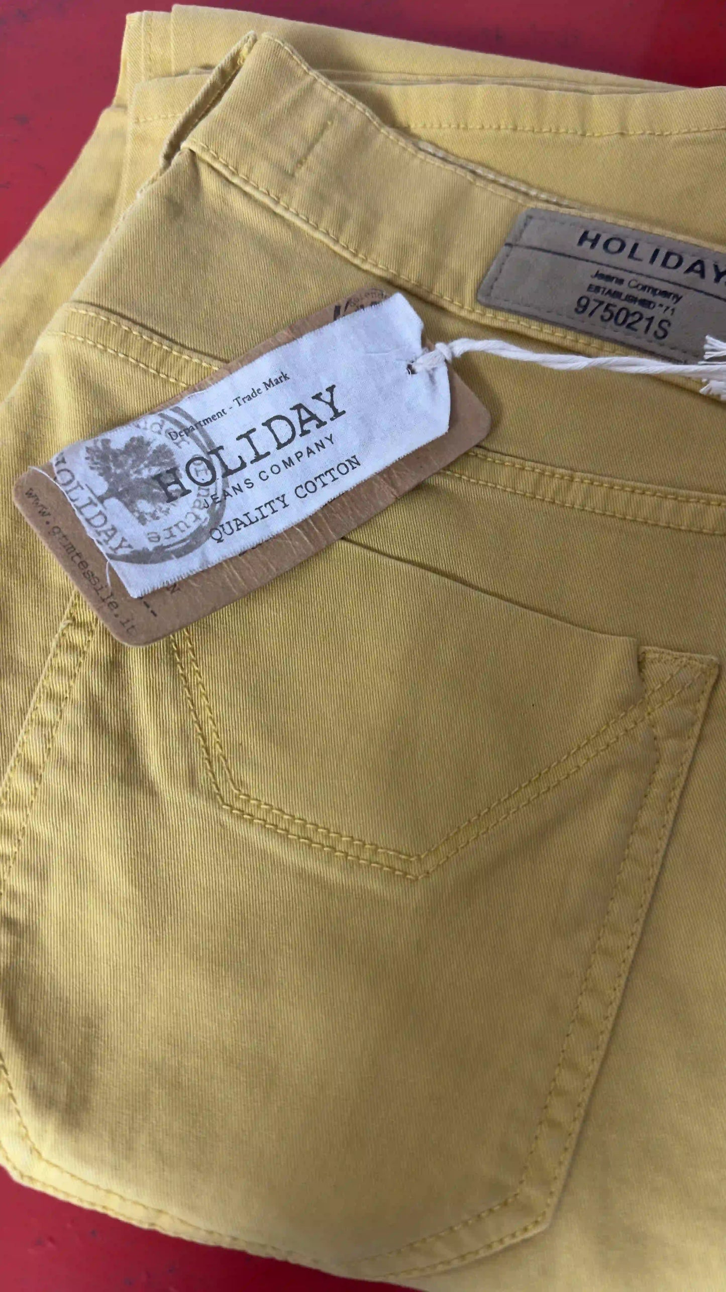 Etichetta Holiday Jeans Company su pantalone ocra piegato su fondo rosso